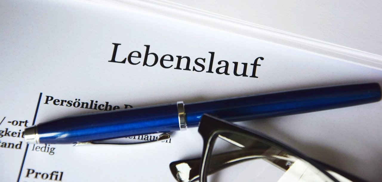 Sie sehen den Schriftzug "Lebenslauf" auf einem Blatt Papier mit mehreren Stiften daneben.